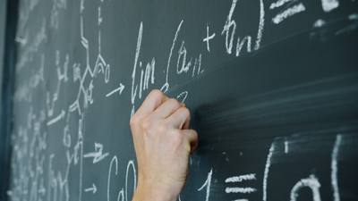 maths student's hand writing formulas with chalk on blackboard indoors in classroom.