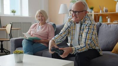 cheerful old man playing video game while woman reading book then smiling and