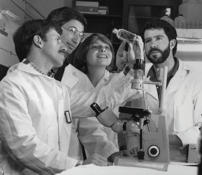 A group of scientists, 1 woman and 3 men, gather around a microscope to examine
