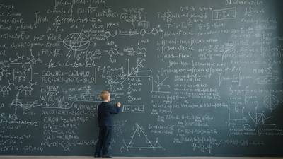 Prodigy child in suit is writing formulas on huge blackboard in school classroom